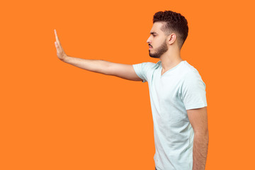 Stop! Side view of serious bossy brunette man in t-shirt standing holding out hand to left,...