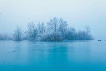 Frozen turquoise lake in misty winter morning, with trees without leaves on a small island across the lake