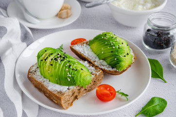 Avocado sandwich on dark rye toast bread made with fresh sliced avocado, cream cheese and seeds on a white plate with a cup of coffee on a light background. Horizontal orientation.