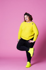 Cheerful plus size model in casual clothes, fat woman in black jeans and yellow jumper on pink...