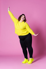 Cheerful plus size model in casual clothes, fat woman in black jeans and yellow jumper on pink...