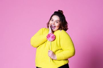 Cheerful plus size model with big lollipop, fat woman in yellow jumper on pink background
