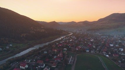 Fototapeta na wymiar pictorial town buildings on winding river bank against hill silhouettes hiding setting sun in orange sky upper view. Carpathian mountains, Ukraine beauty nature. Travel, summer holidays.