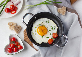 Fried egg with cherry tomato and green onion in frying pan, slices of bread in shape of heart. Tasty romantic breakfast. Top view