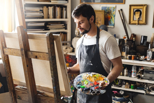 Male painter at art studio indoors holding palette painting on canvas working smiling joyful