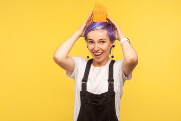 Portrait of nice fashionable hipster girl with violet short hair in denim overalls holding orange paper house on her head and smiling genuinely at camera. studio shot isolated on yellow background
