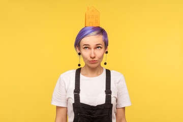 Portrait of cute trendy hipster girl with violet short hair in overalls looking up at orange paper house on her head, thinking over house purchase. indoor studio shot isolated on yellow background