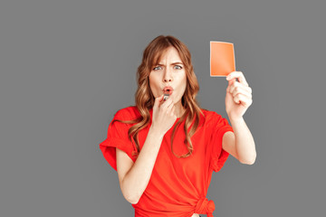 Freestyle. Woman in sports clothes studio standing isolated on grey with red card whistling angry...