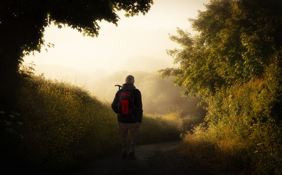 An early morning hiker with a backpack near Newquay in Cornwall. It is very foggy on the trail at the edge of the forest. The sun has risen and shines as backlight.