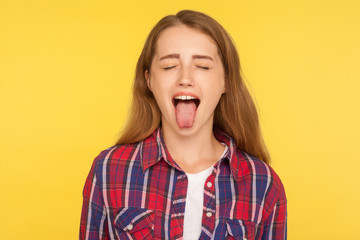 Portrait of naughty disobedient ginger girl in casual shirt standing with closed eyes and sticking out tongue, looking funny childish with derisive grimace. studio shot isolated on yellow background - obrazy, fototapety, plakaty