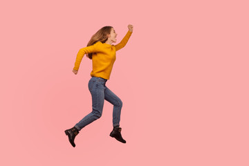 Fototapeta na wymiar Full length portrait of excited ginger girl in sweater and denim jumping in air or flying up, running rushing somewhere, energetic and enthusiasm screaming from happiness. studio shot, pink background