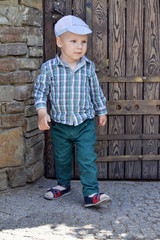 little boy in a shirt, green pants and a cap walks away from a large wooden door