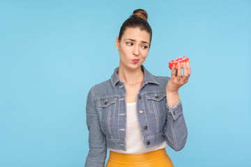 Unhealthy sweet food. Dissatisfied pensive woman with hair bun looking at donut with displeased expression and doubting to eat confectionary, junk food. indoor studio shot isolated on blue background