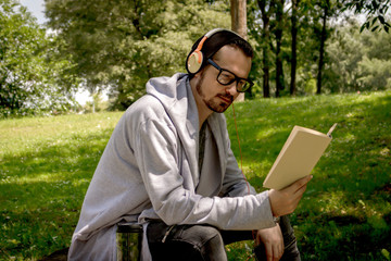Casual man reading book while listening music on headphones in the park.