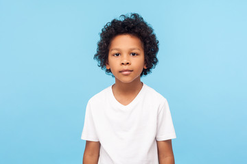 Portrait of cute little boy with stylish curly hairdo in white T-shirt standing, looking at camera...