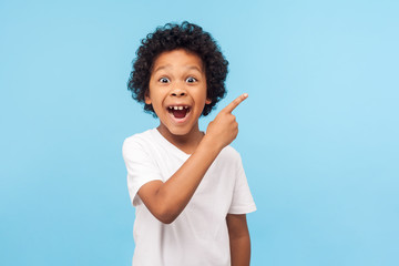 Wow look, advertise here! Portrait of amazed cute little boy with curly hair pointing to empty...