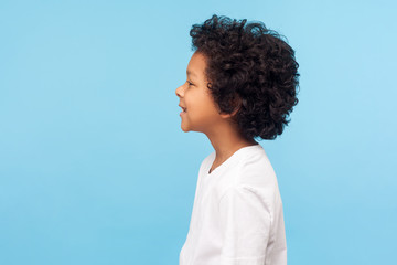 Profile of happy little boy with curly hair in T-shirt smiling carefree and looking to side copy space, healthy cheerful child with positive emotions. indoor studio shot isolated on blue background