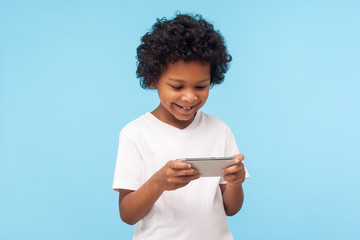 Children entertainment and technology. Happy excited cute little boy with curly hair using mobile phone and smiling, pleased kid playing video game on cellphone, chatting in social network. indoor