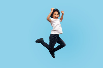 Full length, lively energetic little boy in T-shirt and denim jumping in air screaming with happiness, child flying up, feeling inspired crazy and overjoyed. indoor studio shot, blue background