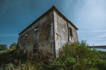 A wide-angle view of a desolate dilapidated stone building in a small European district Alcochete, Portugal, overgrown with bushes, with empty window openings, peeling stucco, the clear sky behind