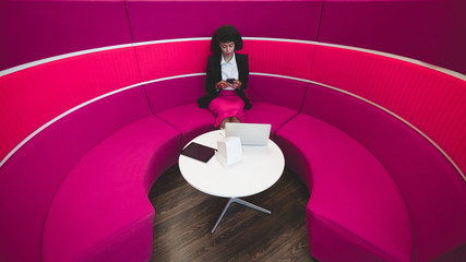 African-American business lady is using her smartphone while sitting on a purple curved sofa in a chillout zone of a modern office; a laptop, charging dock station and a digital tablet on the table