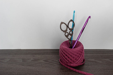 A ball of pink knitted yarn on the table. In the ball are vintage scissors and two hooks (blue and purple). White background