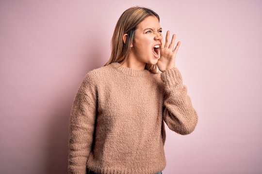 Young beautiful blonde woman wearing winter wool sweater over pink isolated background shouting and screaming loud to side with hand on mouth. Communication concept.