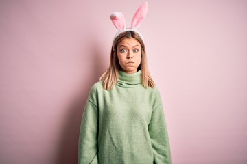 Young beautiful woman wearing easter rabbit ears standing over isolated pink background puffing...