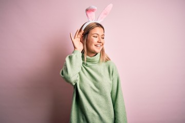 Obraz na płótnie Canvas Young beautiful woman wearing easter rabbit ears standing over isolated pink background smiling with hand over ear listening an hearing to rumor or gossip. Deafness concept.