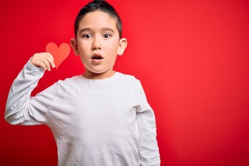 Young little boy kid holding heart paper shape over isolated red background scared in shock with a surprise face, afraid and excited with fear expression