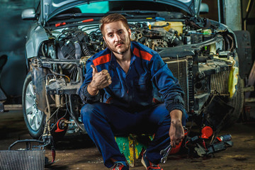 Plakat A tired mechanic in a blue protective suit is sitting near a disassembled car. Repair Service Concept.