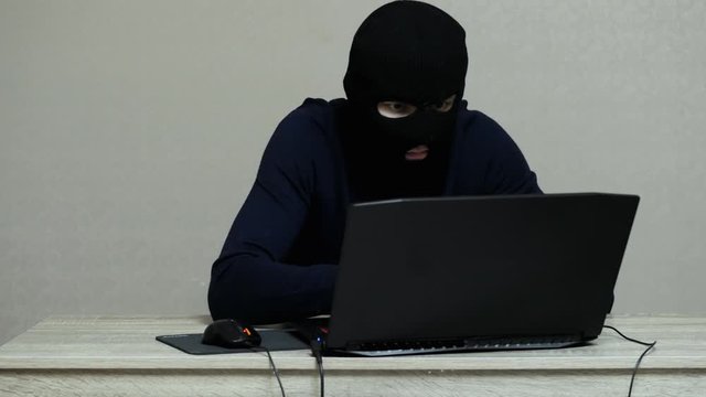 Funny male hacker in black mask balaclava do hacker attack on laptop, looking at the camera and thumbs up
