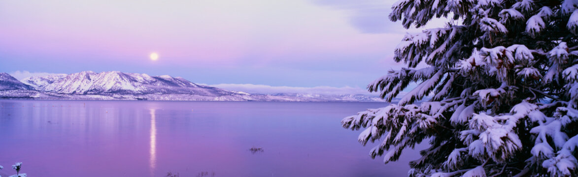 This is Lake Tahoe after a winter snow storm. There is a full moon rising over the lake and the trees and mountains are covered in snow.