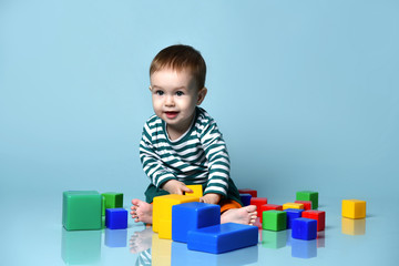 Obraz premium Little baby boy in stylish casual clothing barefoot sitting on floor and playing with colorful toy cubes over blue wall background