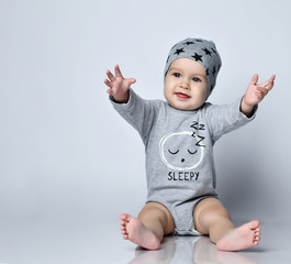 Little baby boy toddler in grey casual jumpsuit, cap and barefoot sitting on floor and smiling with...