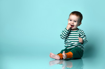 Little baby boy in stylish casual clothing barefoot sitting on floor and smiling over blue wall...