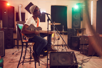 Full length portrait of contemporary African-American man singing to microphone and playing guitar during rehearsal or concert with music band in recording studio, copy space