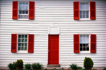 Fototapeta na wymiar This is a close up of a white house with red shutters. The front door is also red. There are a few small bushes in front of the house.