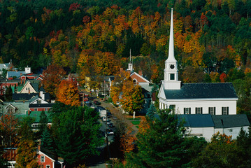 This is Scenic Route 100 in the autumn. There is a large white New England style church with a tall steeple next to smaller buildings of the town.