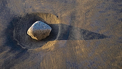 Large irregular boulder in stong side light, casting a long shadow, on a mixed basalt and silica sandy background, a moon landscape concept.