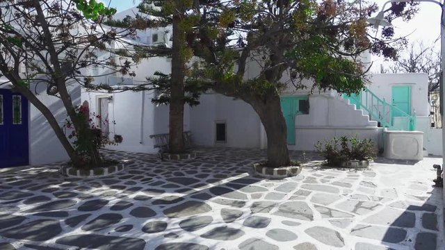Sliding view of an empty, tiled courtyard with trees casting shadows in front of typical white, Greek houses on the island of Mykonos