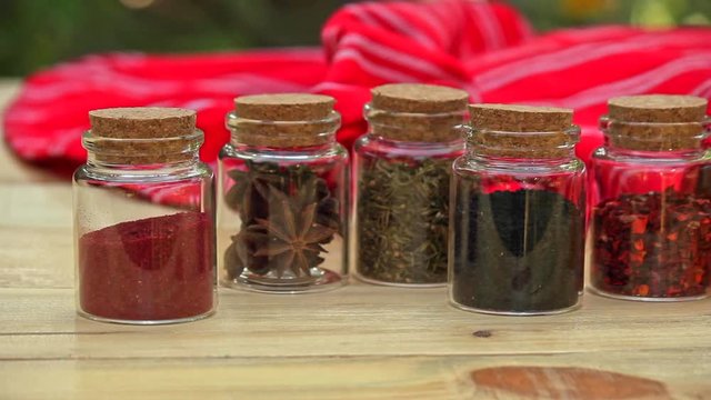 Selection of Jars with dried spices laid out in a row on wooden table with red cloth in the background. slow pan right