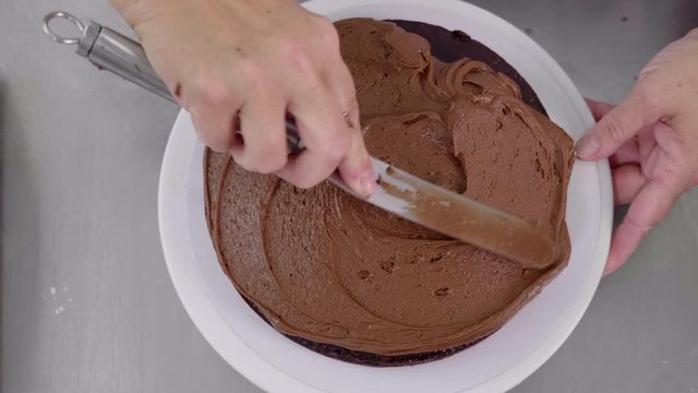Top down shot of chocolate cake being topped with butter cream. The cake is turned on a lazy suzy by a female chef, the buttercream spread with a palette knife. Camera moves in slowly to the cake.