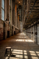 Stool sitting in the hallway of an abandoned prison, with shadows on the floor from the bars in the...