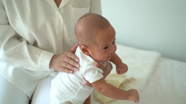 From above of mom patting adorable newborn son massaging back of Asian baby on bed at home