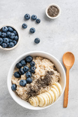 Fototapeta na wymiar Oatmeal porridge bowl with banana slices, chia seeds and blueberries on grey concrete backdrop, top view with copy space, vertical composition