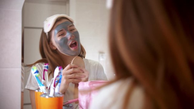 A young attractive woman with a mask on her face dancing and singing in front of the mirror in bathroom
