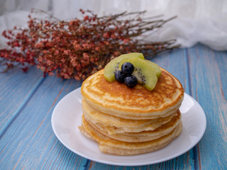 Stack of pancakes with topping, kiwi and blueberry.placed in a white plate on a blue wooden table.Eat with milk tea in the glass.