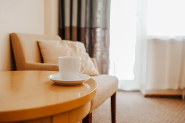 Fototapeta na wymiar white cup on a wooden coffee table against the background of a beige armchair with a pillow, elegant interior in the style of minimalism, a place for relaxation or business meetings