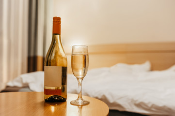 bottle of alcoholic drink and a beautiful glass with white wine and bubbles, on a wooden bedside table against the background of dim light and a blurry bed